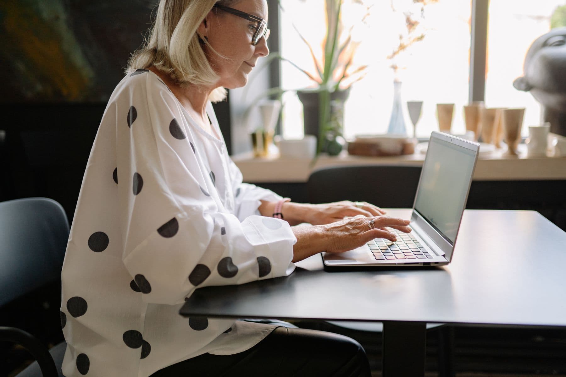A woman typing on a laptop at a table by a bright window